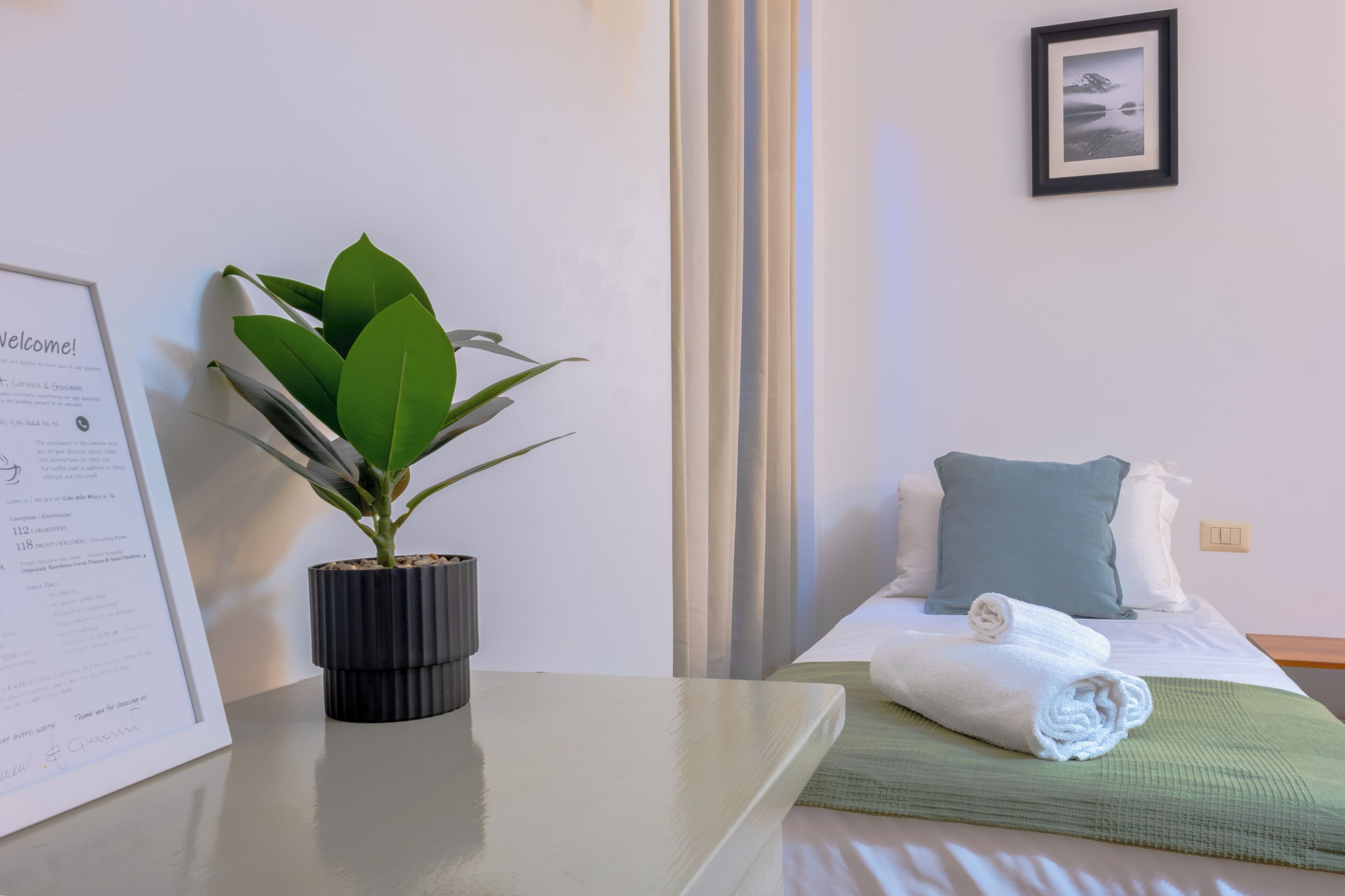 A hotel room with a bed, a rolled towel, a green throw, a potted plant, and a welcome sign.