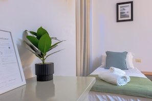 A hotel room with a bed, a rolled towel, a green throw, a potted plant, and a welcome sign.