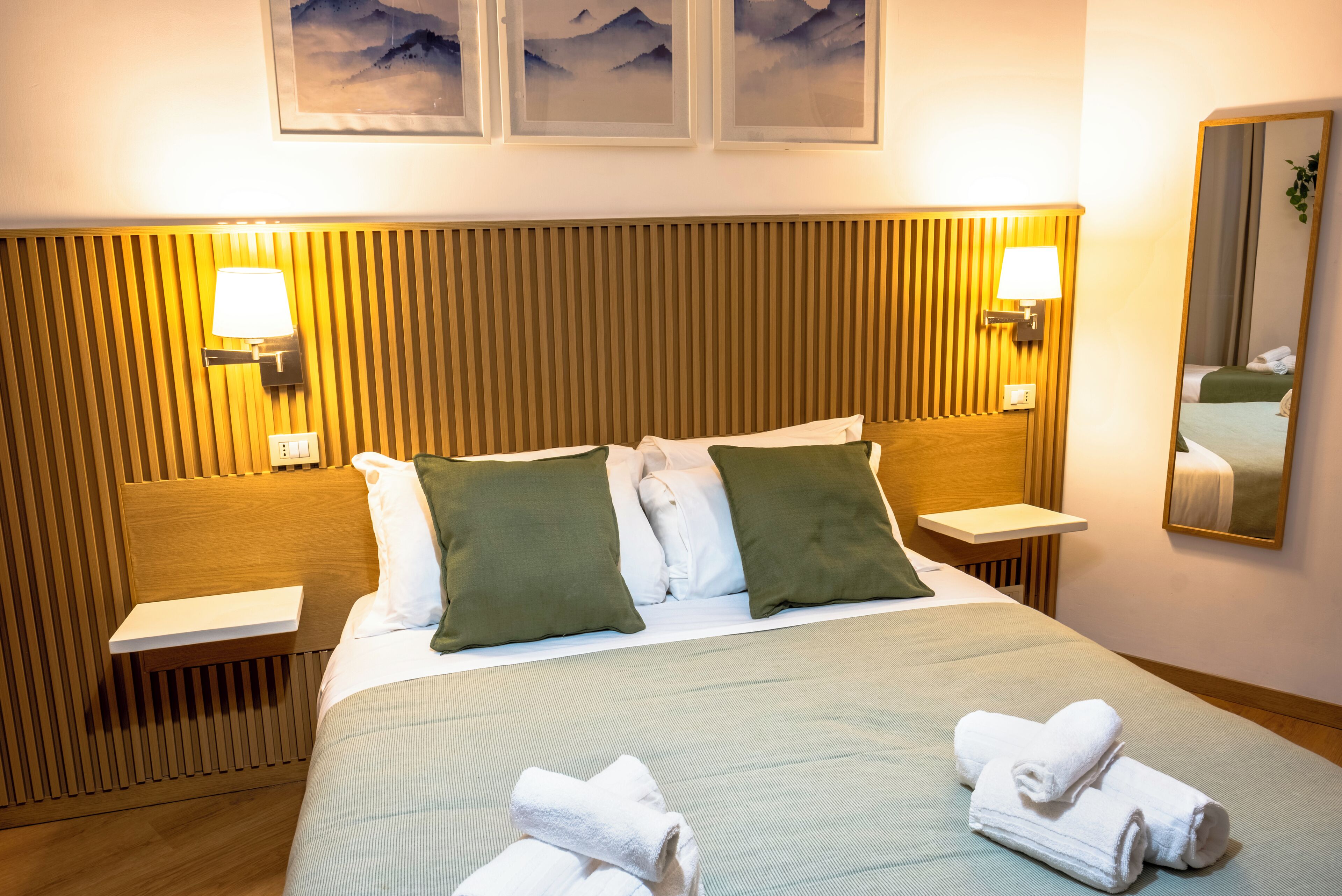 Habitació d'hotel amb un capçal de llit de fusta, dues taules de nit, un llit amb roba de llit blanca i almoines verdes, una làmpada penjada a la paret i un mirall.