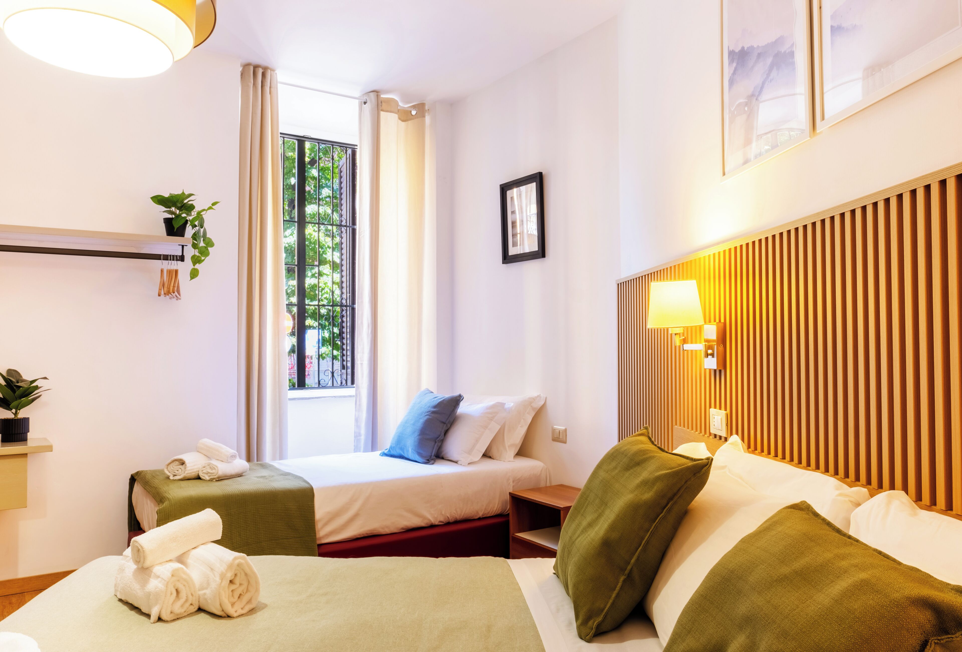 A hotel room with two beds, a window with curtains, a bedside table with a lamp, and a wall-mounted shelf with a plant.