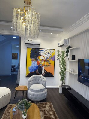 A modern living room with a chandelier, a large abstract painting, a grey armchair, a flat-screen TV, and an air conditioning unit. - Luxury 4-Bedroom Duplex in Lekki with Snooker, Jacuzzi, Fast Wi-Fi & 24/7 Power (Lekki)