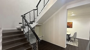 A staircase with a black railing leading to an upper floor, a white wall with a picture, and a dining area with a table and chairs. - Luxury 4-Bedroom Duplex in Lekki with Snooker, Jacuzzi, Fast Wi-Fi & 24/7 Power (Lekki)