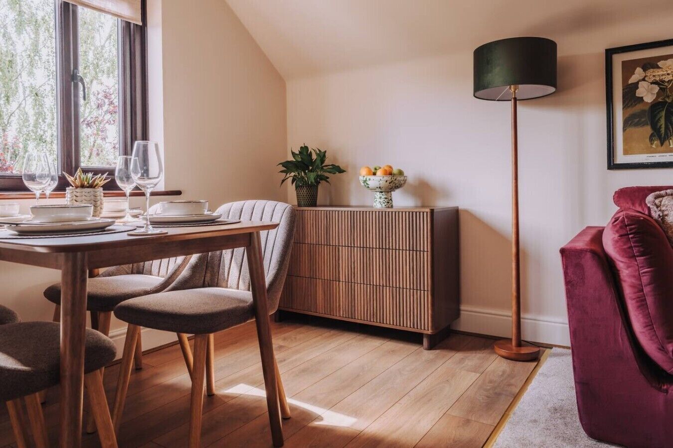 A dining area with a wooden table set for two, a cabinet with a plant, and a red sofa.
