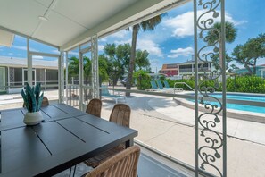 Überdachte Terrasse mit Tisch und Stühlen, mit Blick auf einen Swimmingpool und Palmen.