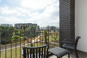 A balcony with two chairs, a view of a landscaped area with a water feature, and modern buildings in the background. - Park Place | 1 Bed with Balcony and Lake Views (Cape Town)