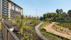 Ein modernes Gebäude mit mehreren Balkonen, eine angelegte Grünfläche mit einem gewundenen Holzweg und ein Gewässer.