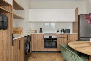 A compact kitchen with wooden cabinets, a Bosch oven, a washing machine, and a dining area with a green upholstered chair. - Park Place | 1 Bed with Balcony and Lake Views (Cape Town)