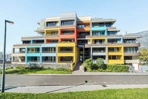 Un edificio de varios pisos con balcones y ventanas amplias, ubicado en una zona verde bajo un cielo despejado.