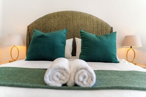 Ein ordentlich bezogenes Bett mit einem grünen Kopfteil, zwei türkisfarbenen Kissen und einem Paar zusammengerollter Handtücher.