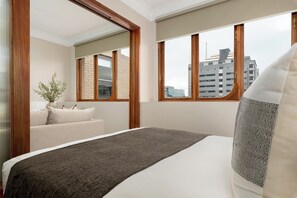 A hotel room with a bed, a window with a view of buildings, a plant, and a wooden door. - Charlie and the Chairman | Elegant Urban Stay (Cape Town)
