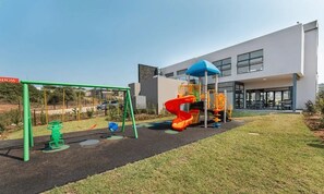 A playground with swings and a slide, a building with a blue roof, and a grassy area. - Warm and Cosy Apartment | Ballito Village (Dolphin Coast)