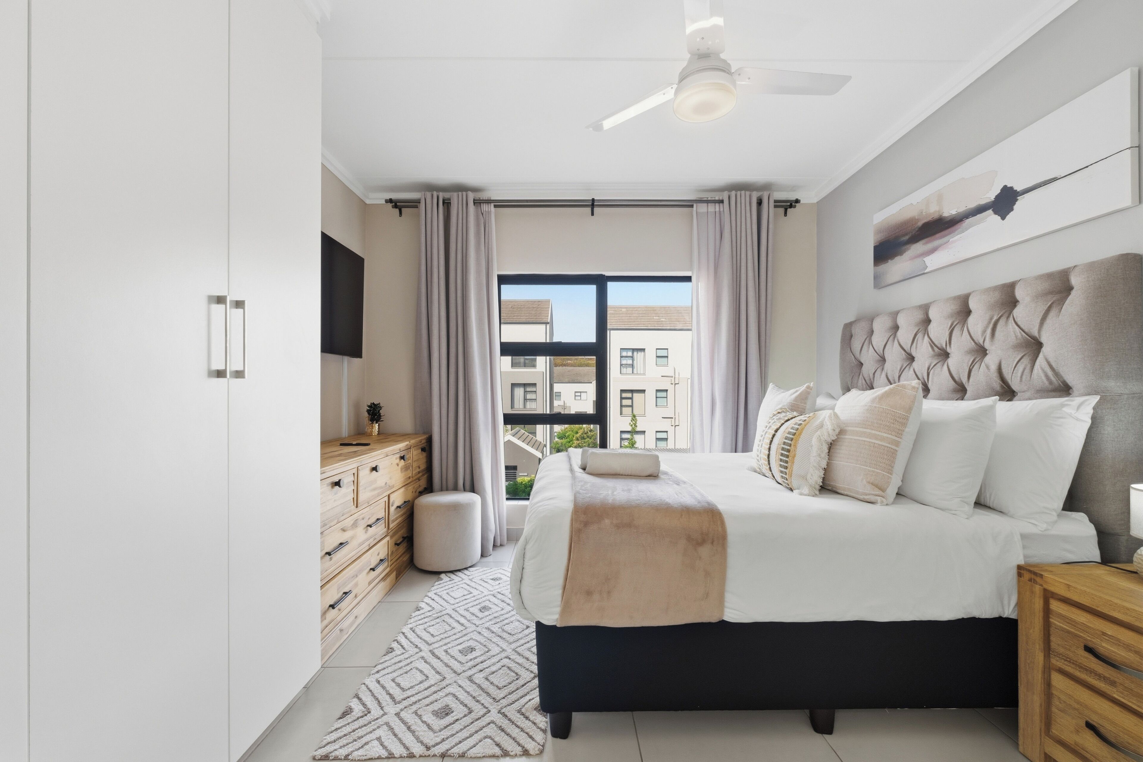 Een moderne slaapkamer met een groot bed, een plafondventilator, een televisie en uitzicht op gebouwen.