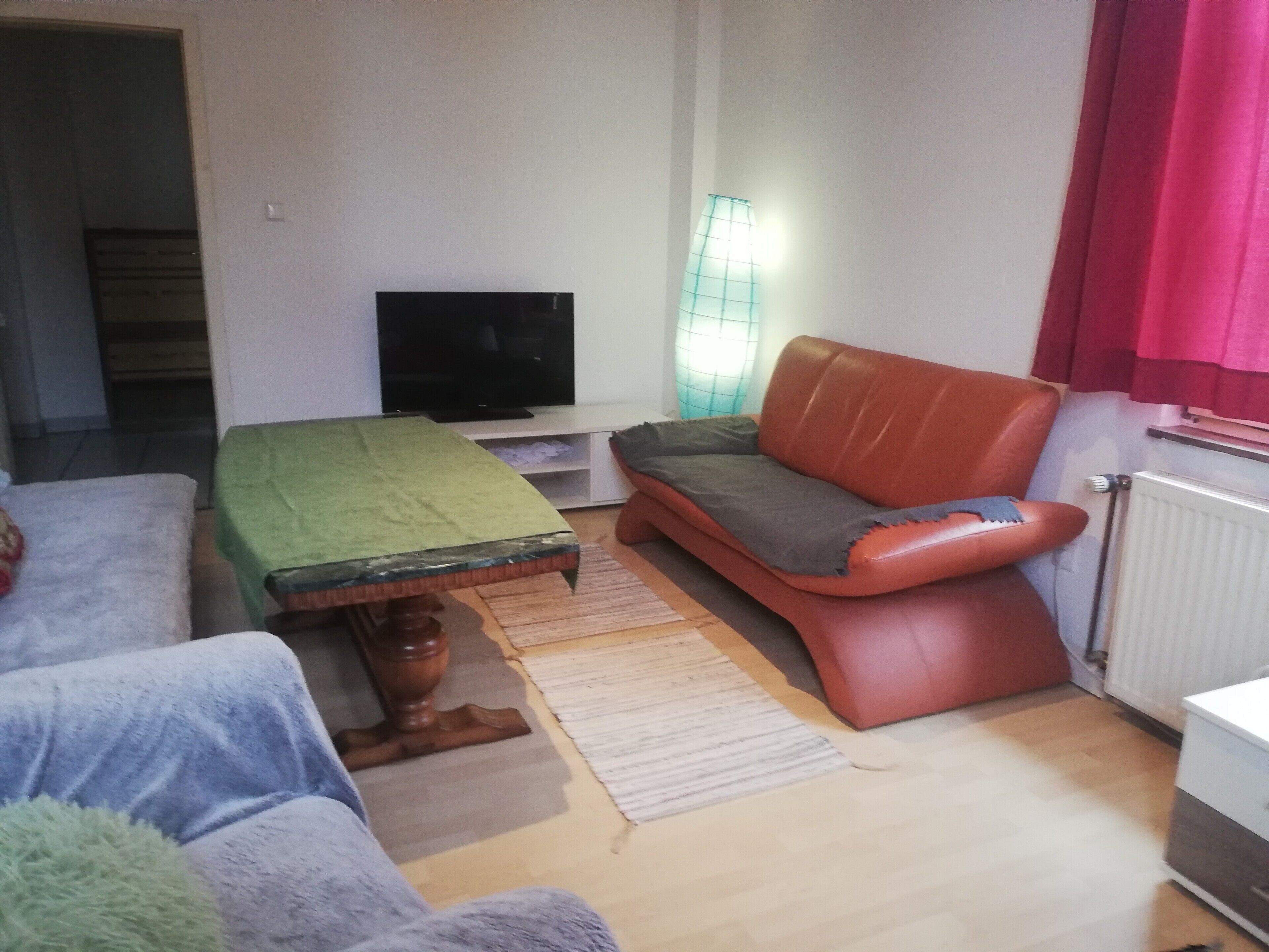 A living room with a sofa, a television, a table with a green tablecloth, and a radiator.
