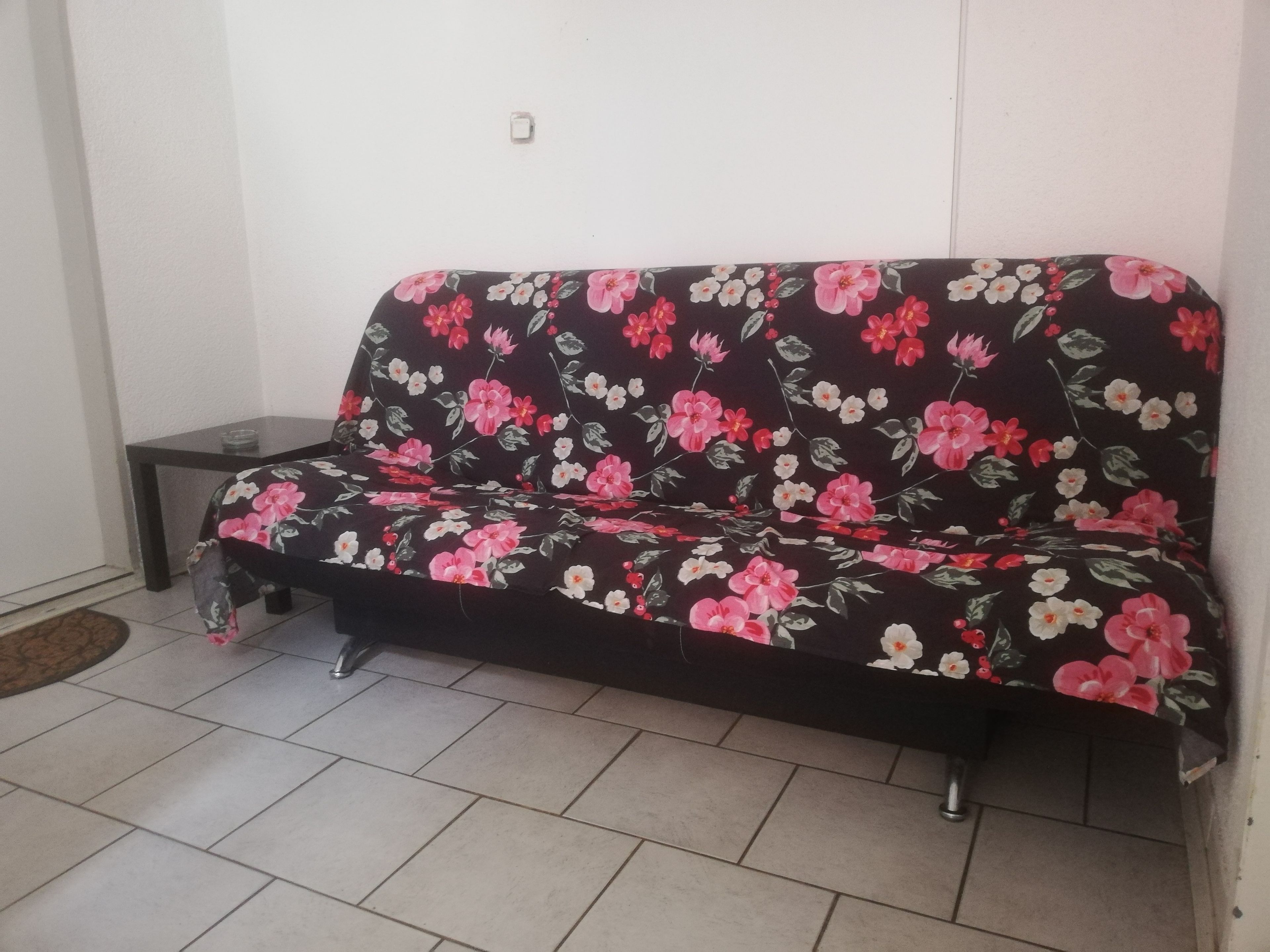A dark-colored sofa with a floral pattern, positioned against a white wall in a room with tiled flooring.