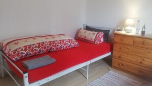 En seng med en rød madras, et tæppe med blomstermønster og puder. En trækommode med en lampe og en flaske ovenpå.