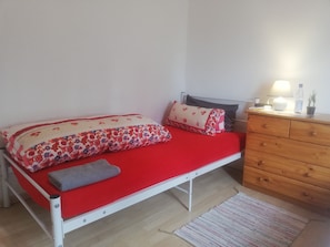 A bed with a red mattress, a floral-patterned blanket, and pillows. There is a wooden dresser with a lamp and a bottle on top.