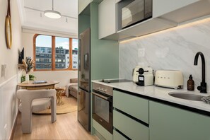 Eine kompakte Küche mit Einbauofen, Mikrowelle und modernen Geräten. Ein Essbereich mit kleinem Tisch und Stuhl ist zu sehen, und ein Fenster bietet Blick auf die Stadtlandschaft.