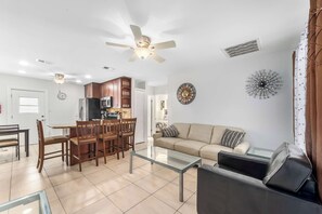 A modern living room with a glass coffee table, a sofa, and a dining area with wooden chairs and a kitchen in the background. - 3 Min Walk to Beach | Private Triplex Retreat (Indian Rocks Beach)