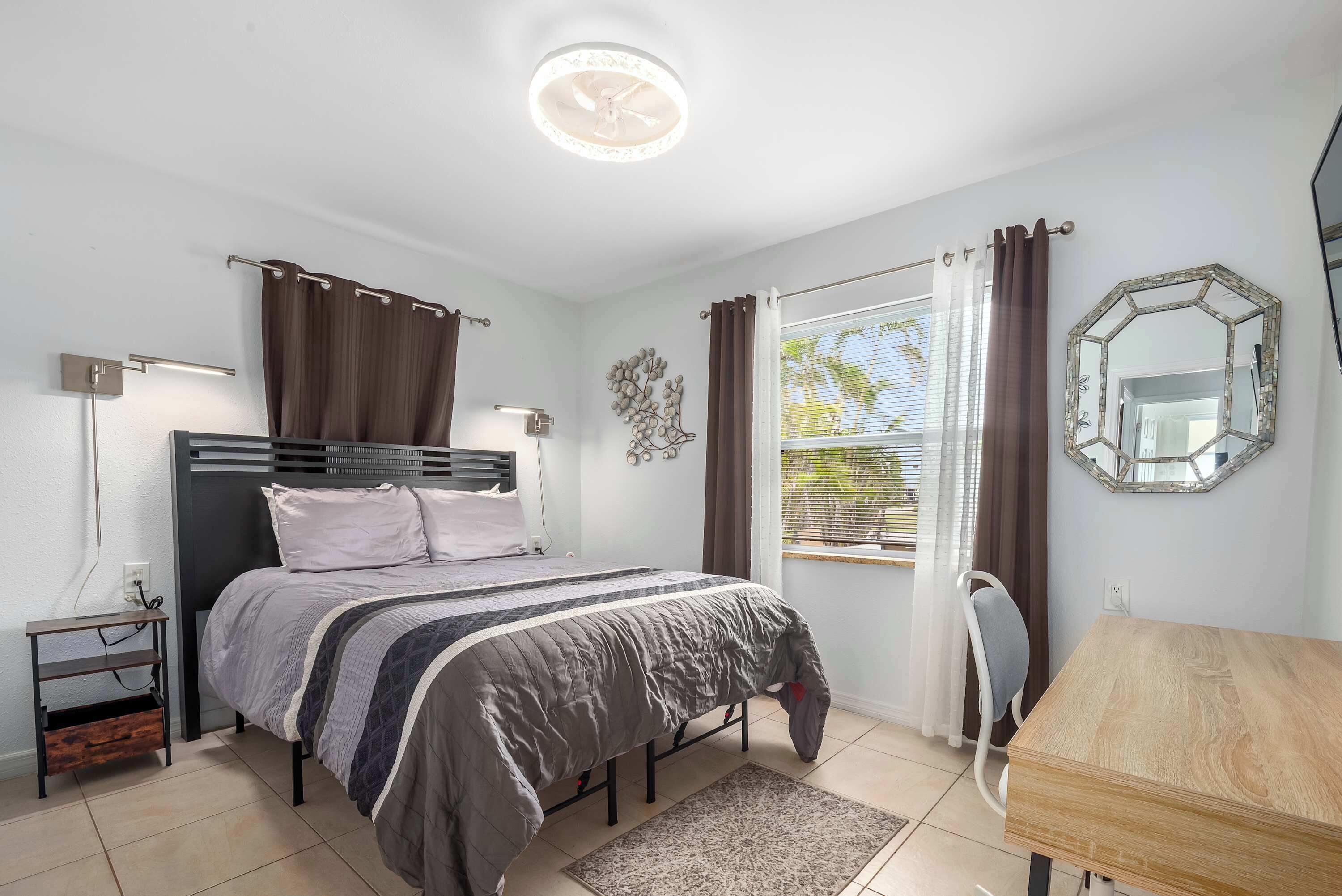 Ein Schlafzimmer mit einem Bett, Nachttischen, einem Schreibtisch, einem Stuhl, einem Fenster mit Vorhängen, einem Wandspiegel und einer Deckenleuchte.