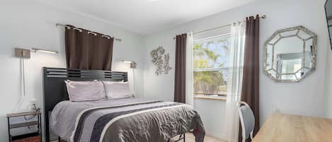 A bedroom with a bed, bedside tables, a desk, a chair, a window with curtains, a wall-mounted mirror, and a ceiling light.