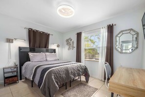 Quarto com cama, mesas de cabeceira, escrivaninha, cadeira, janela com cortinas, espelho fixado na parede e luminária no teto.