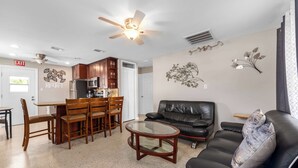 A living room with a black leather sofa, a glass coffee table, wooden dining chairs, a kitchen area with cabinets, a ceiling fan, and wall art. - 3 Min Walk to Beach | Private Triplex Retreat (Indian Rocks Beach)