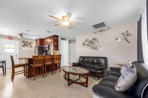 Ein Wohnzimmer mit schwarzem Ledersofa, einem Glastisch, Holz-Esessstühlen, einer Küchenzeile, einem Deckenventilator und Wandbildern.