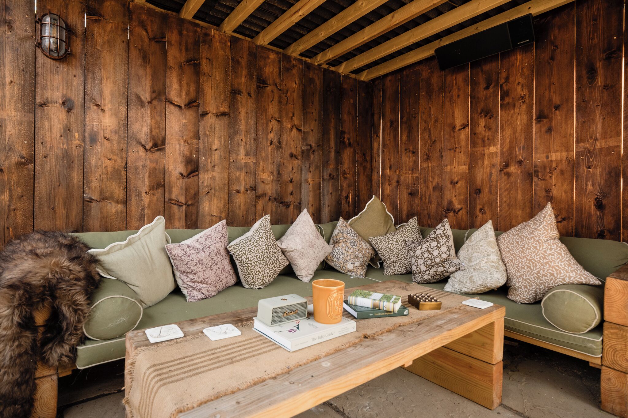 A wooden interior with a sofa and cushions, a coffee table with books and a mug, and a lamp.