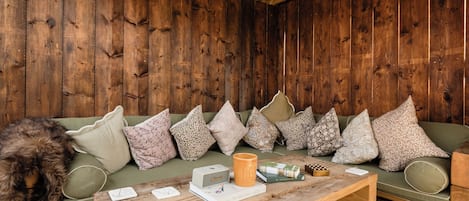 A wooden interior with a sofa and cushions, a coffee table with books and a mug, and a lamp.