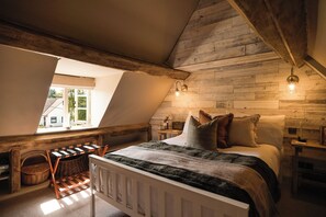 Ein Schlafzimmer mit schrägem Dach, einem Bett mit Kissen, einem Nachttisch, einer Lampe und einem Fenster mit Blick ins Grüne.