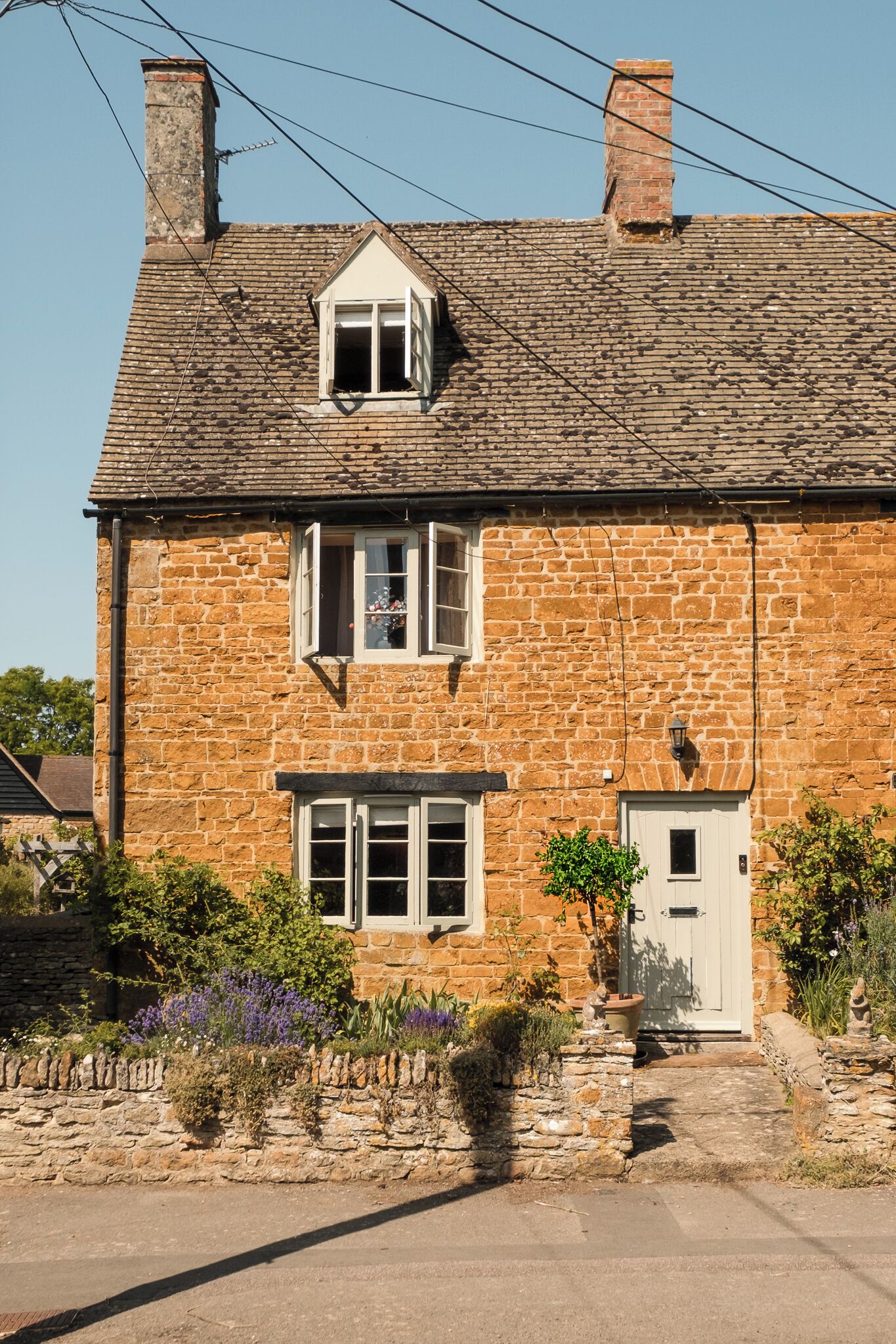 Une maison en pierre avec un toit à pignon, deux fenêtres, une porte et un petit jardin fleuri.
