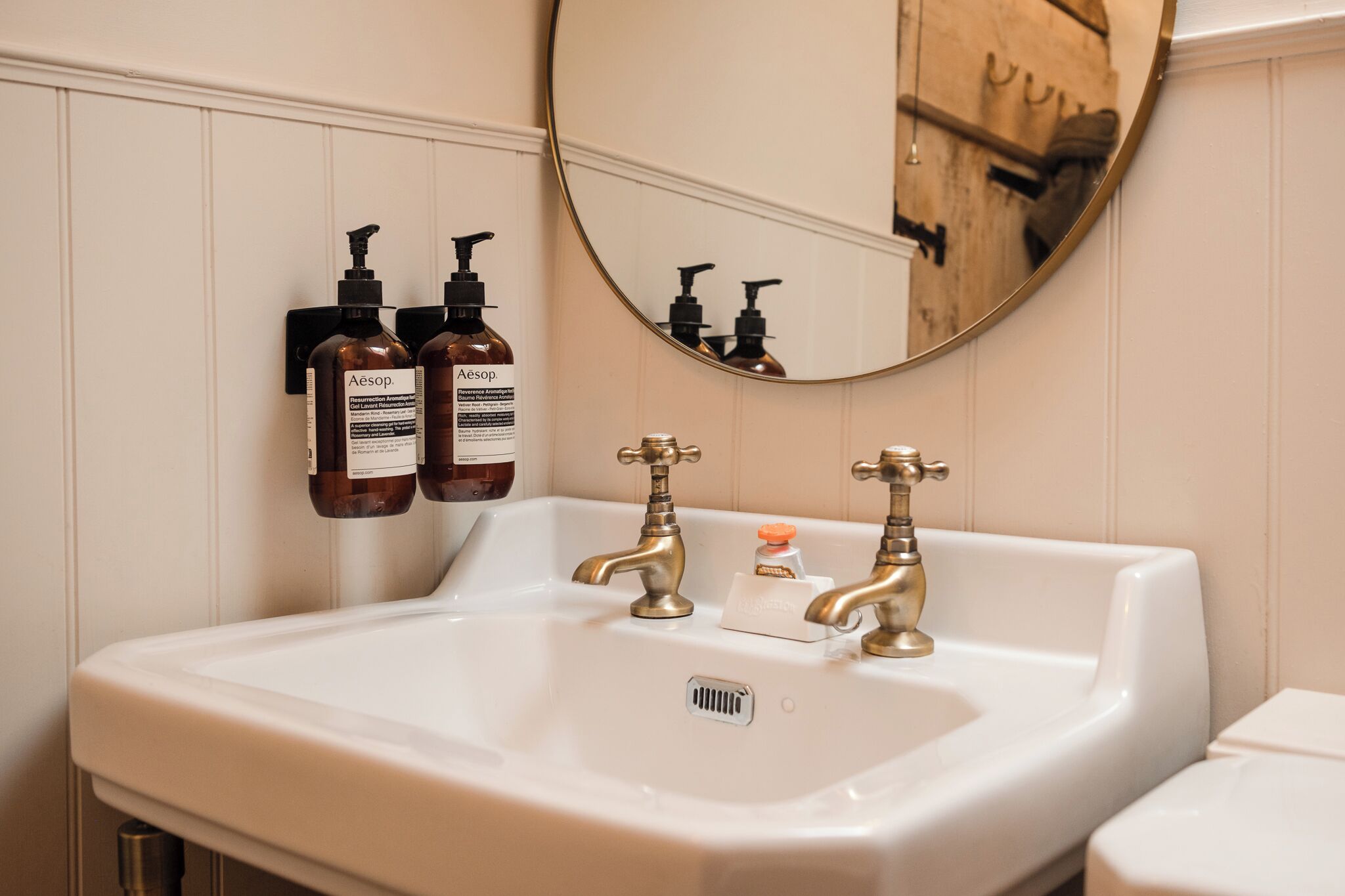 Un baño con un lavamanos blanco, dos grifos dorados y dos botellas de productos Aesop en la pared.