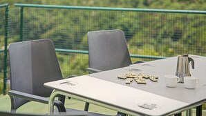 Una mesa con una cafetera, tazas y fichas de dominó, frente a una cerca y follaje verde.