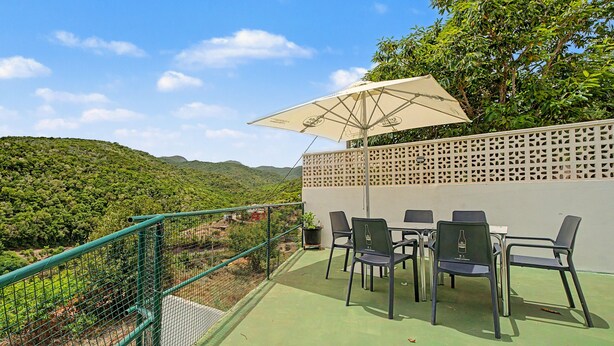 An outdoor seating area with a table and chairs, a green fence, and a view of a hilly landscape.