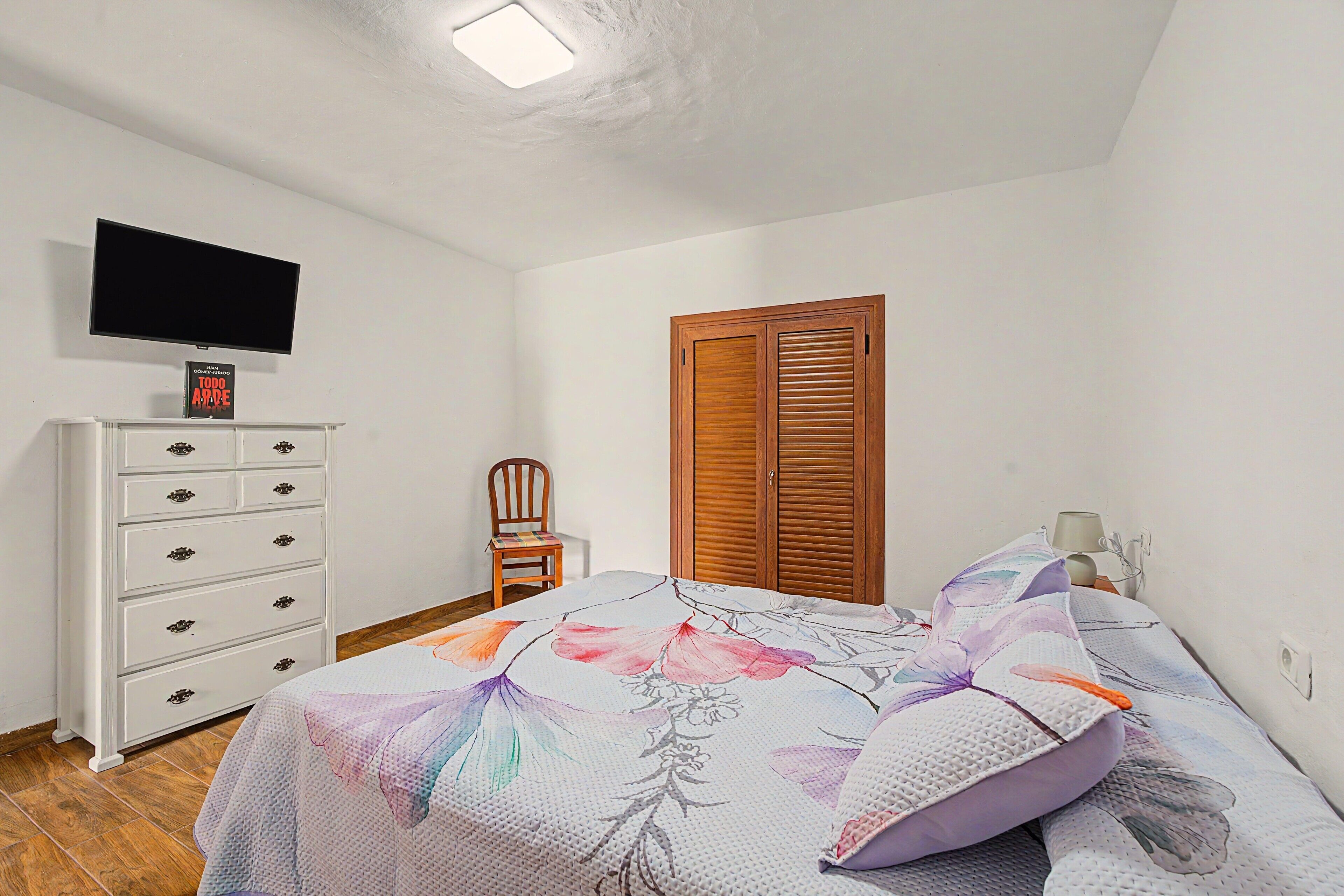 A bedroom with a bed, a television on the wall, a wooden wardrobe, a chair, and a bedside table with a lamp.
