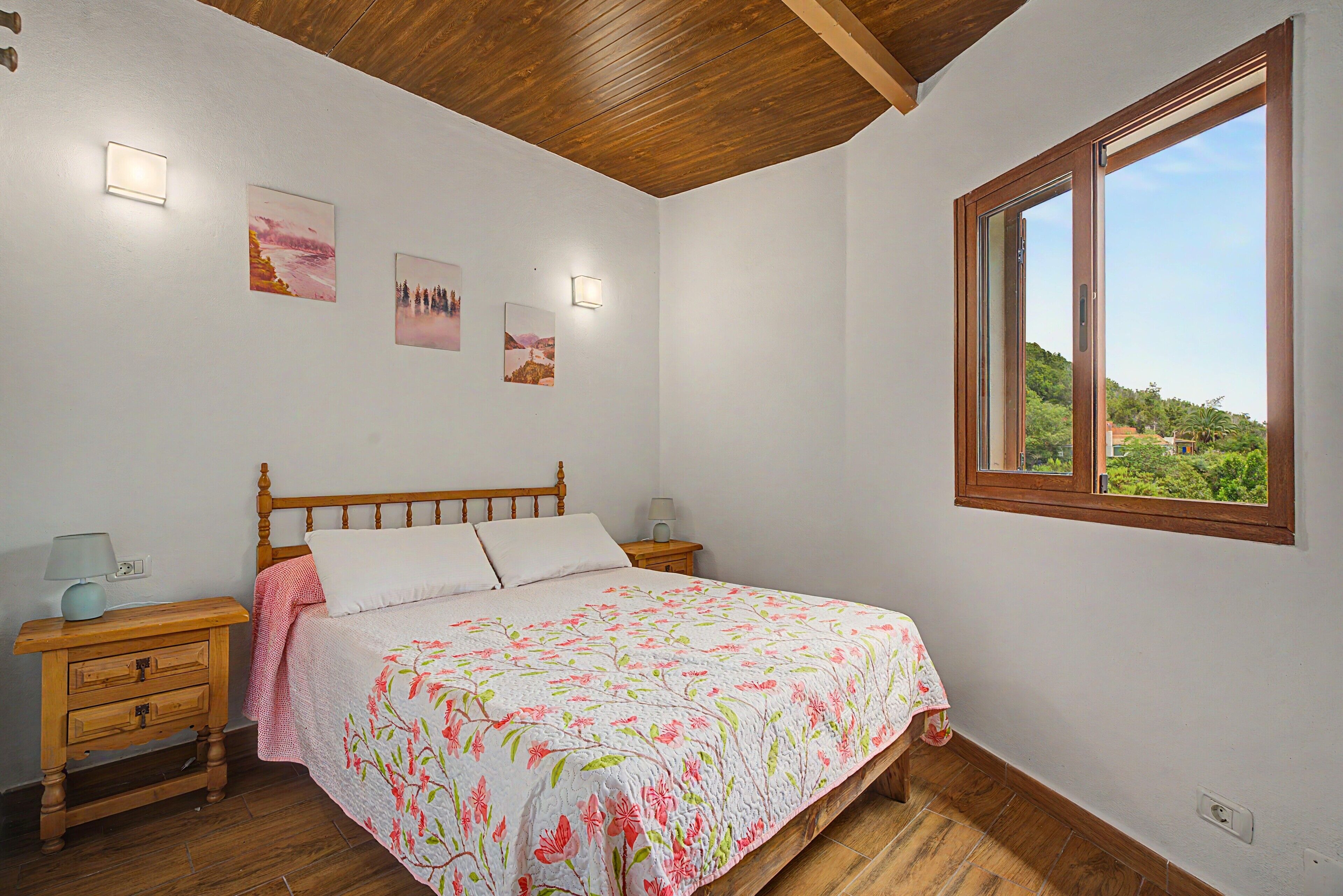 A bedroom with a wooden bed, a nightstand, a lamp, a window with a view, and wall-mounted lights.