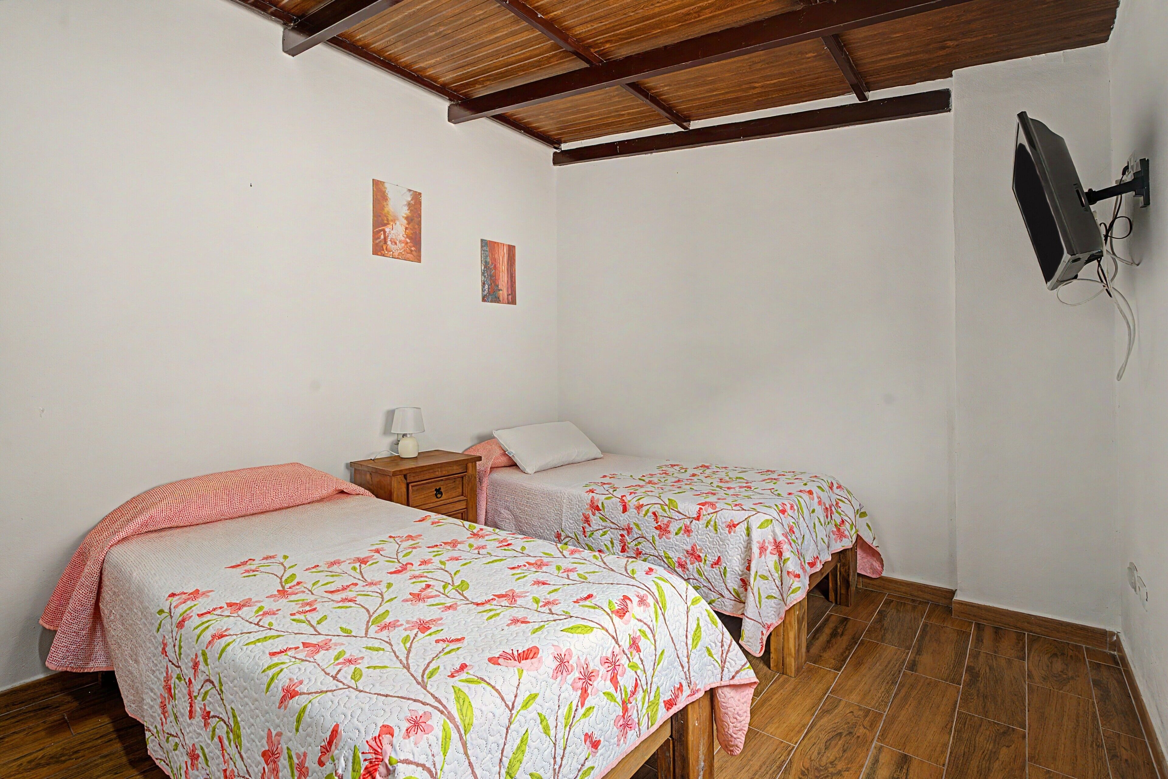 Quarto com duas camas de solteiro, piso de madeira, televisão fixada na parede e uma pequena mesa de cabeceira com abajur.