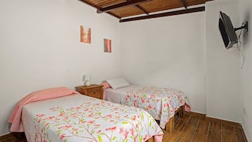 Quarto com duas camas de solteiro, piso de madeira, televisão fixada na parede e uma pequena mesa de cabeceira com abajur.