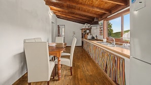 Una cocina con vigas de madera en el techo, una zona de comedor con mesa y sillas, y vista al exterior a través de una ventana.