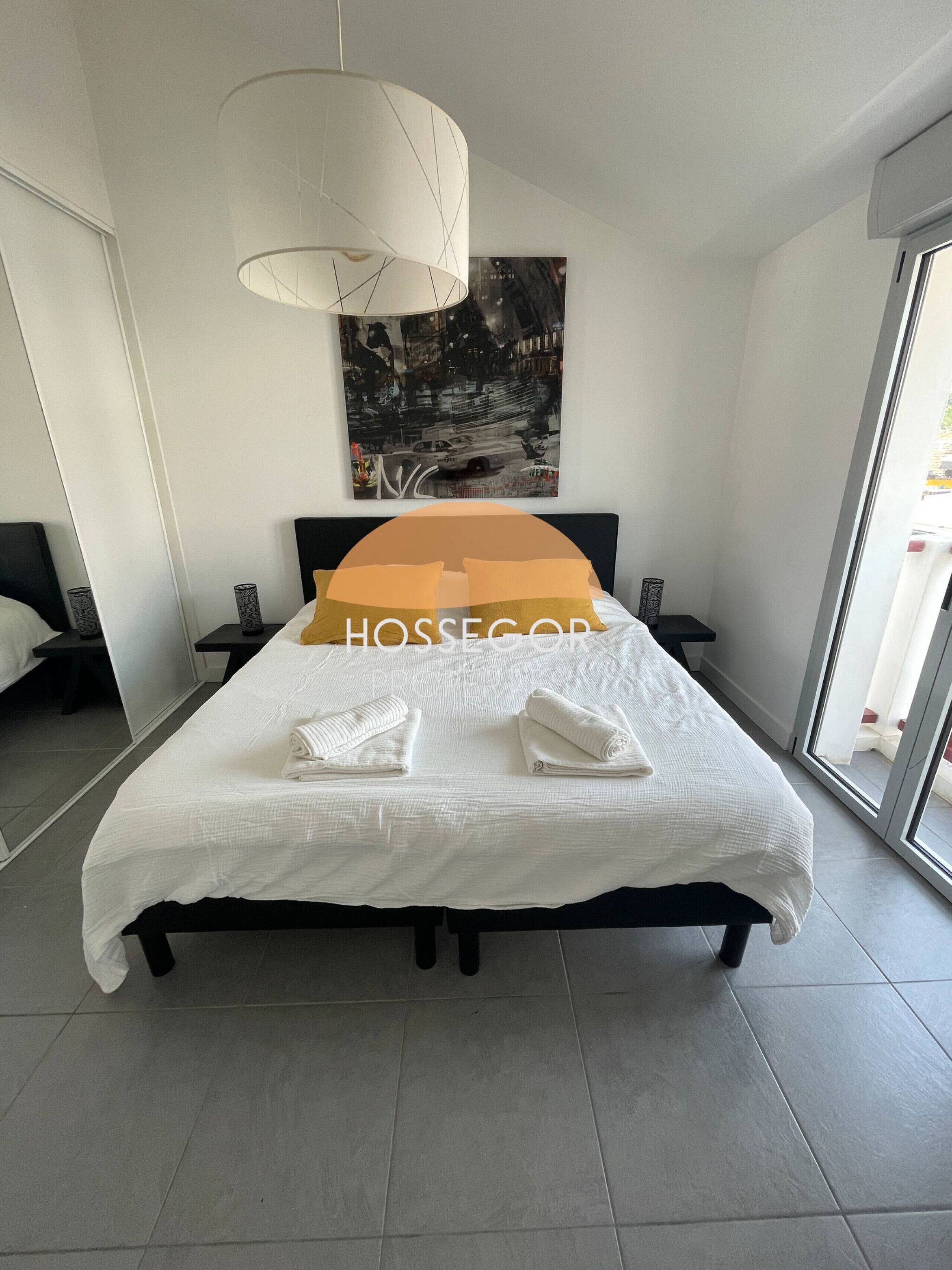 Une chambre moderne avec un grand lit, un luminaire suspendu, un tableau représentant un paysage urbain et une vue à travers une porte vitrée.
