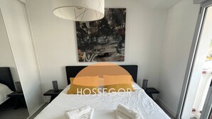 Ett modernt sovrum med en stor säng, en hängande lampa, en målning som föreställer en stadsbild och en utsikt genom en glasdörr.