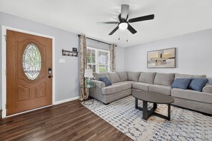A living room with a wooden door, a ceiling fan, a grey sectional sofa, a coffee table, and a patterned rug.
