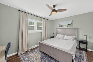 A bedroom with a bed, a nightstand, a ceiling fan, a window with curtains, and a painting on the wall.