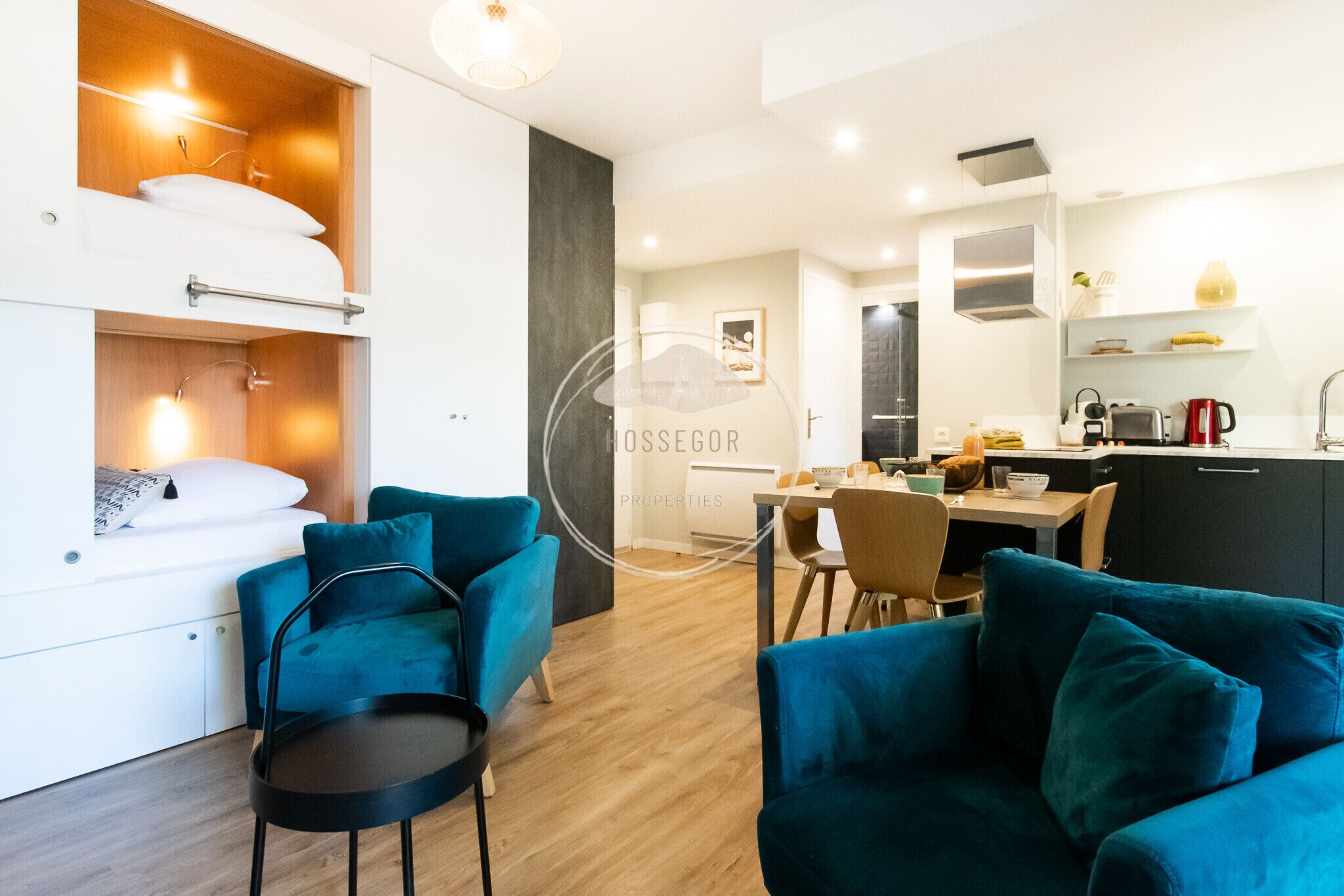 Ein modernes Wohnzimmer mit Türkis-Farbenen Sofas, ein Essbereich mit Tisch und Stühlen sowie eine Küche mit zeitgemäßen Geräten.