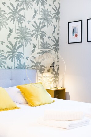 A bedroom with a bed, pillows, a nightstand, a lamp, and a wall with a tropical wallpaper. - Appt Ermitage - Hossegor center - Terrace - Park (Soorts-Hossegor)