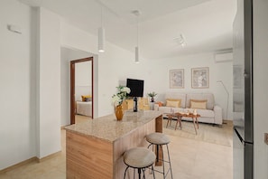 A modern kitchen with a central island, bar stools, a flat-screen TV, and a sofa in the background.