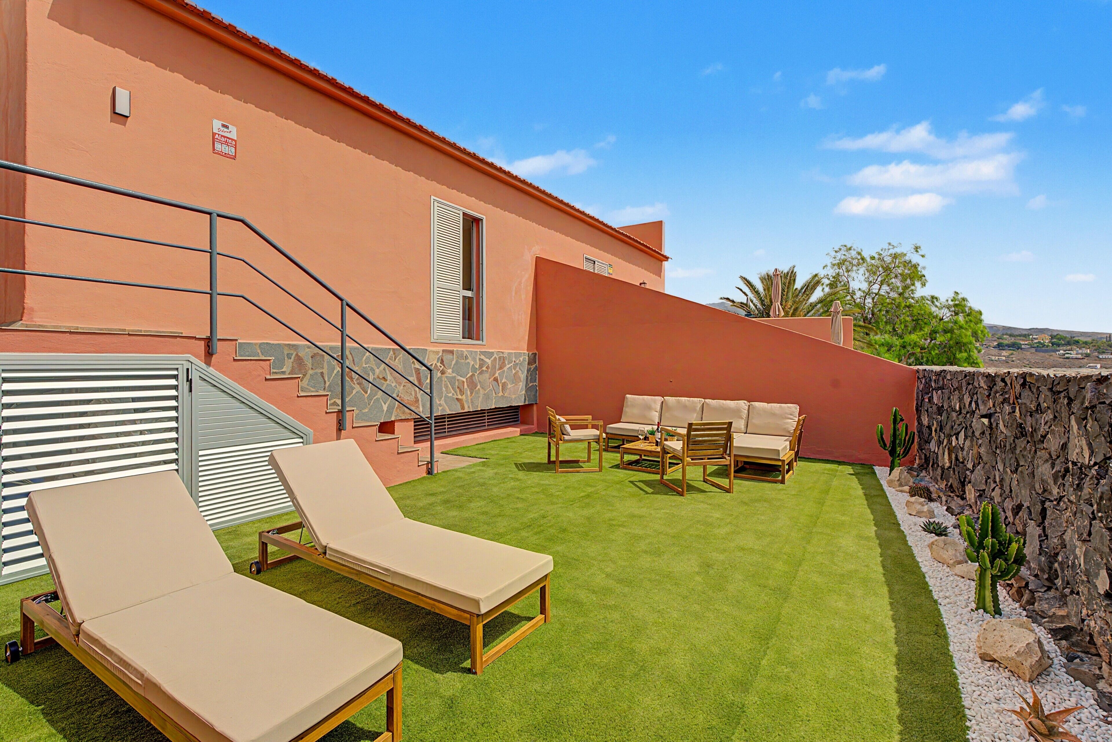 A patio with two lounge chairs, a stone wall, and a staircase leading to a second floor.