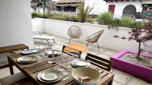 A wooden dining table set with plates, glasses, and a bowl, surrounded by wicker chairs, on a patio with a view of buildings and a small tree. - Appt Brémontier 3 - Hossegor center - Terrace (Soorts-Hossegor)