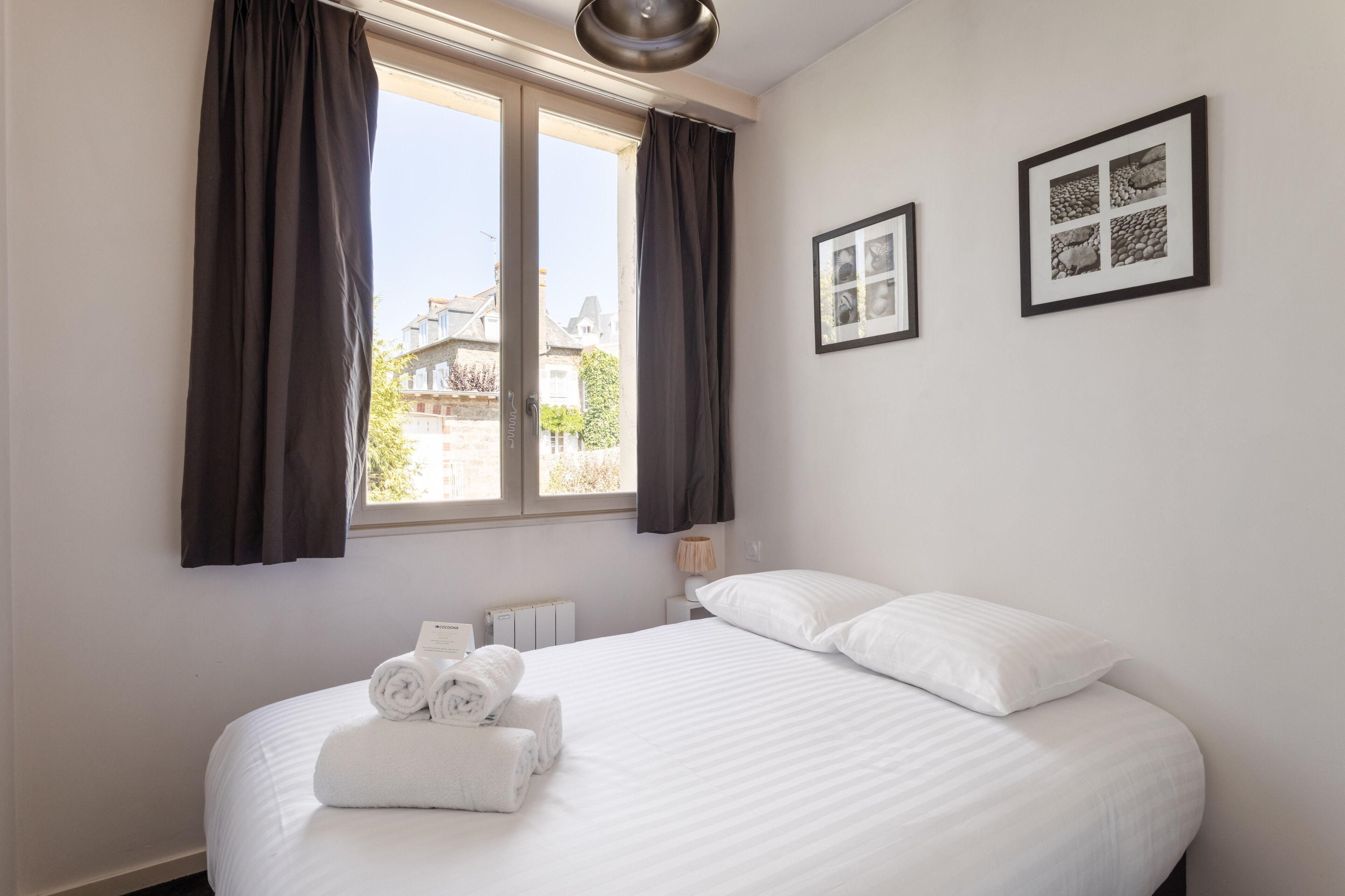 Habitación de hotel con una cama grande, dos cuadros en la pared, una ventana con cortinas y un radiador.