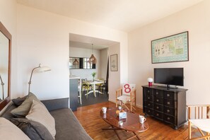 Ein Wohnzimmer mit einer Couch, einem Couchtisch mit Tassen, einem Fernseher auf einer Kommode und einem Essbereich im Hintergrund.